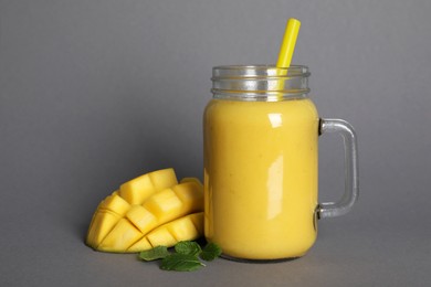 Photo of Mason jar of tasty smoothie with straw, mango and mint leaves on grey background
