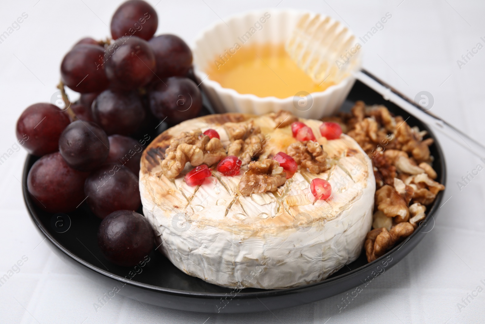 Photo of Plate with tasty baked camembert, honey, grapes, walnuts and pomegranate seeds on white tiled table, closeup