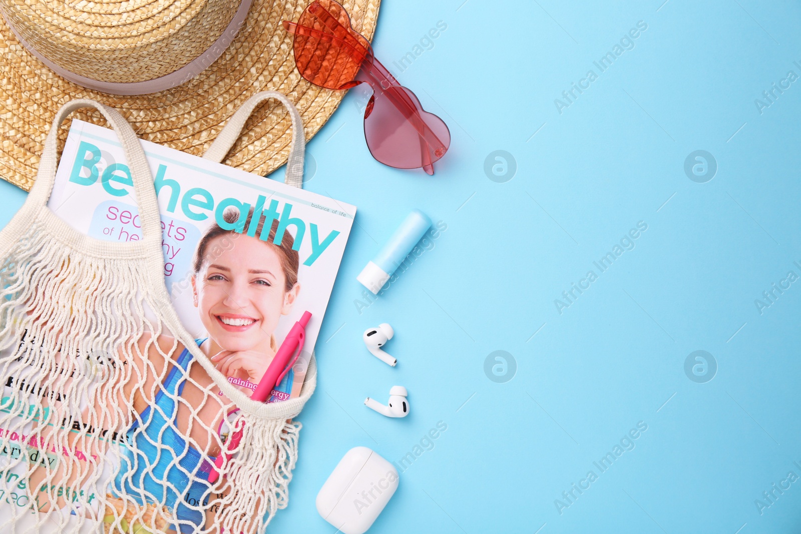 Photo of String bag with magazine, earphones and summer accessories on light blue background, flat lay. Space for text