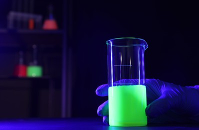 Photo of Scientist with laboratory beaker of luminous liquid at table against dark background, closeup. Space for text