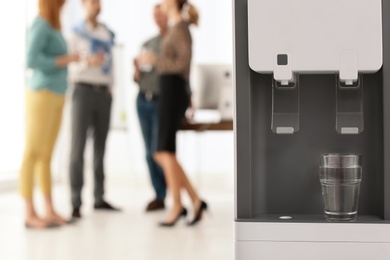 Photo of Modern water cooler with glass and blurred office employees on background. Space for text