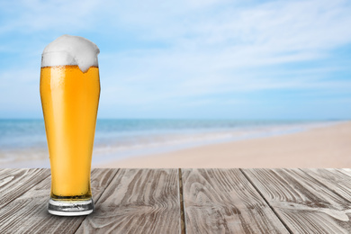 Image of Cold beer on wooden desk against blurred sea and sandy beach. Space for text