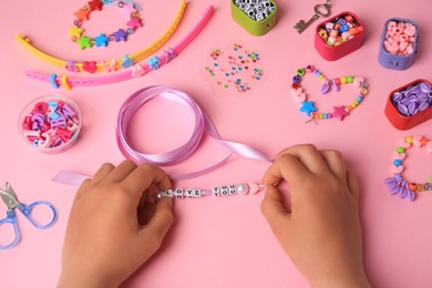 Photo of Child making beaded jewelry and different supplies on pink background, above view. Handmade accessories