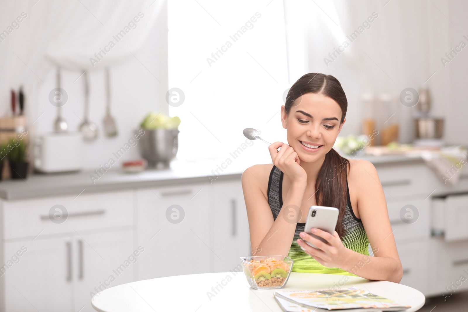 Photo of Young woman in fitness clothes using smartphone while having healthy breakfast at home. Space for text