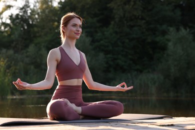 Photo of Beautiful woman practicing Padmasana on yoga mat on wooden pier near pond, space for text. Lotus pose
