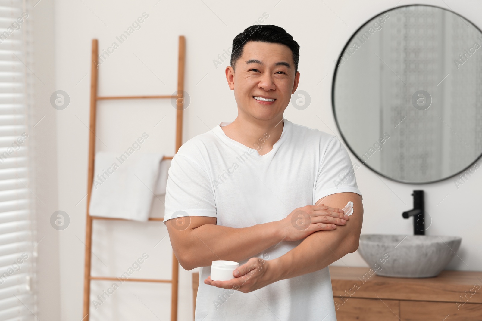 Photo of Handsome man applying body cream onto his arm in bathroom