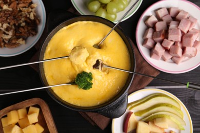 Photo of Dipping different products into fondue pot with melted cheese on black wooden table, flat lay