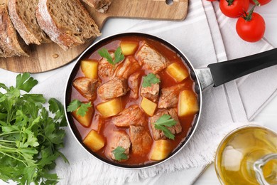 Photo of Delicious goulash in saucepan, bread and ingredients on white table, flat lay