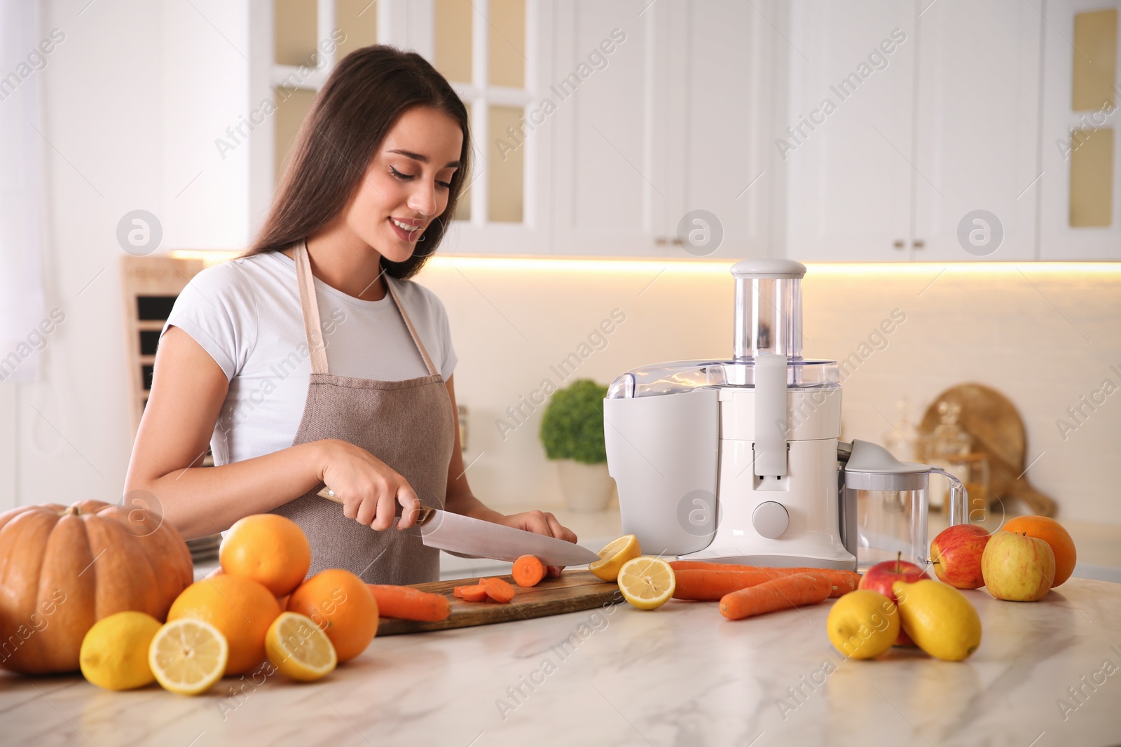 Photo of Young woman cutting fresh carrot for juice at table in kitchen