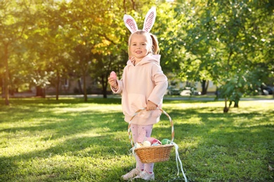 Cute little girl with bunny ears and basket of Easter eggs in park