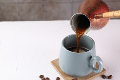 Photo of Pouring aromatic coffee from cezve into cup at white textured table, closeup. Space for text