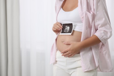 Pregnant woman with ultrasound picture of baby near window indoors, closeup. Space for text