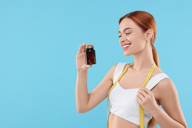 Photo of Happy young woman with bottle of pills and measuring tape on light blue background, space for text. Weight loss