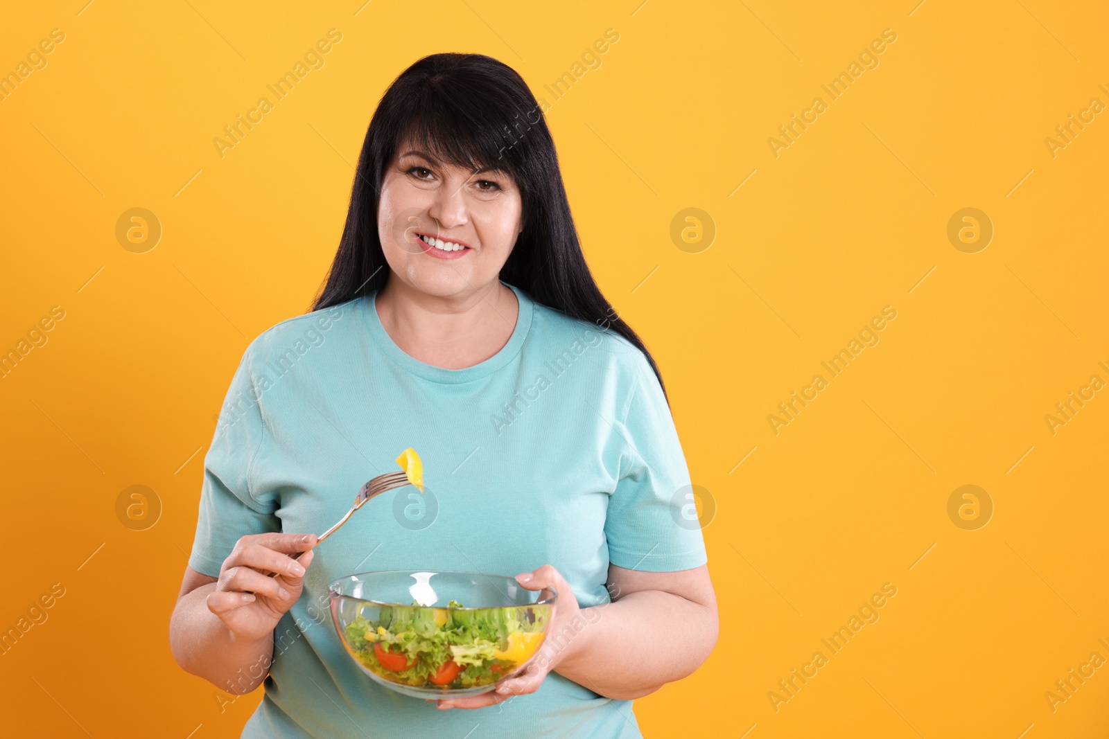 Photo of Beautiful overweight woman eating salad on yellow background, space for text. Healthy diet