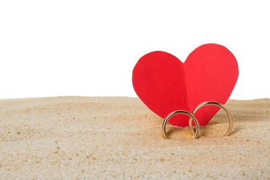 Honeymoon concept. Two golden rings, red paper heart and sand isolated on white