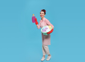 Photo of Housewife holding bottle of cleaning product and basin with clothes on light blue background