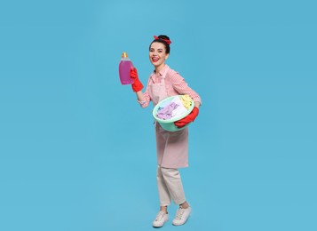 Housewife holding bottle of cleaning product and basin with clothes on light blue background