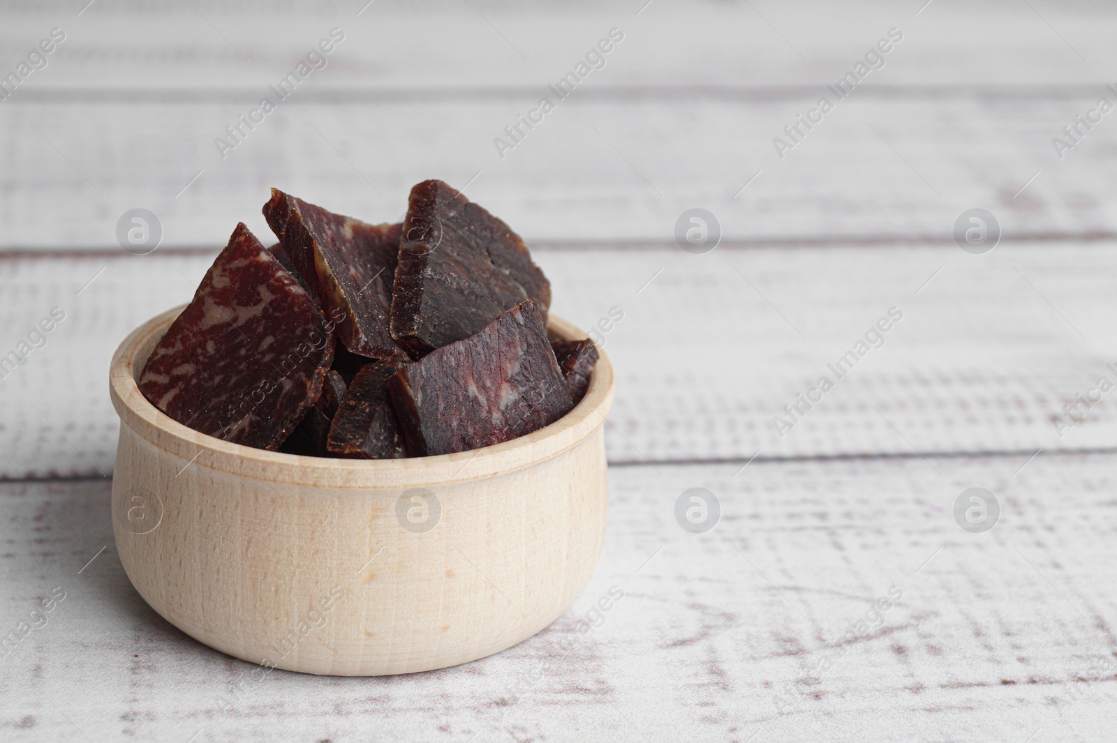 Photo of Delicious beef jerky in bowl on white wooden table, space for text