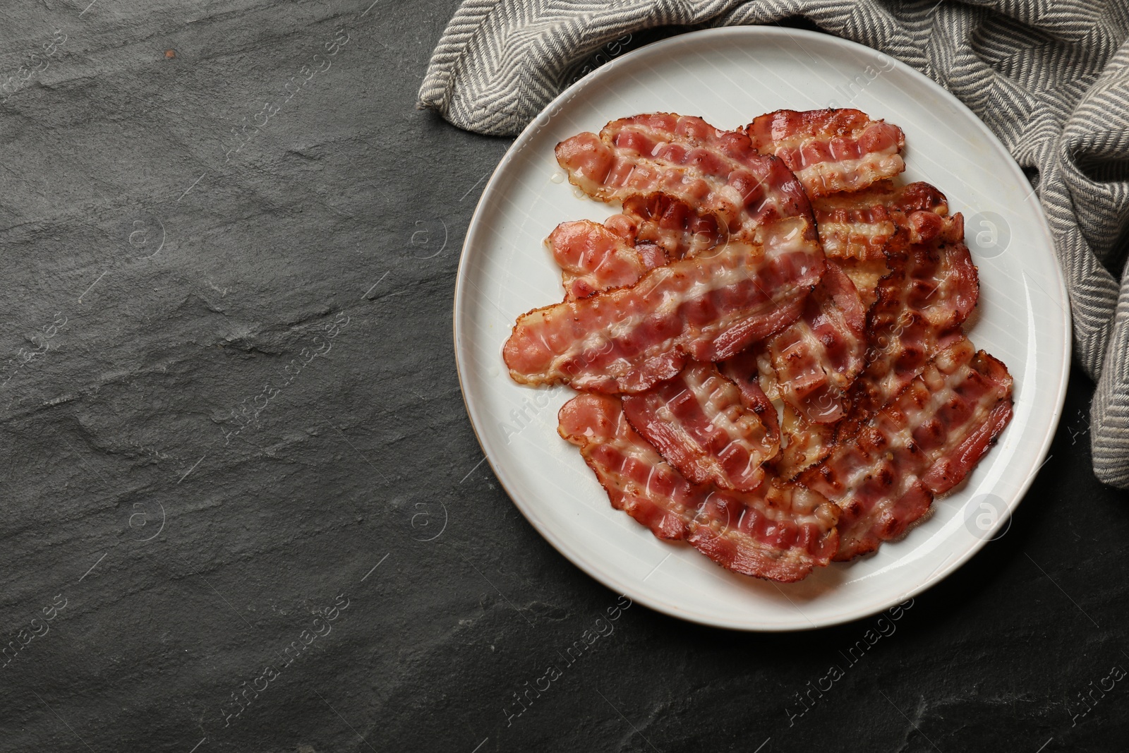 Photo of Plate with fried bacon slices on dark textured table, top view. Space for text