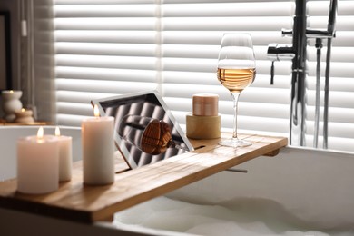 Photo of Wooden tray with tablet, wine and candles on bathtub in bathroom