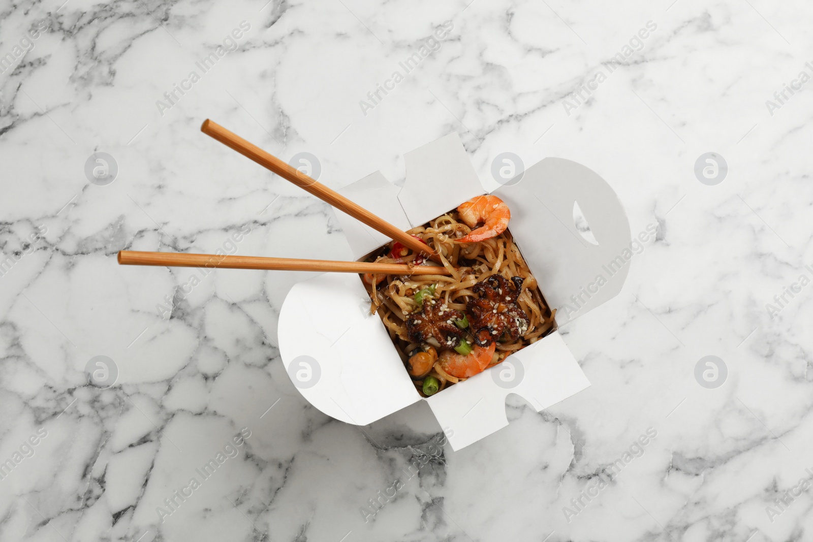 Photo of Box of wok noodles with seafood and chopsticks on white marble table, top view