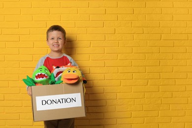 Cute little boy holding donation box with soft toys near yellow brick wall, space for text