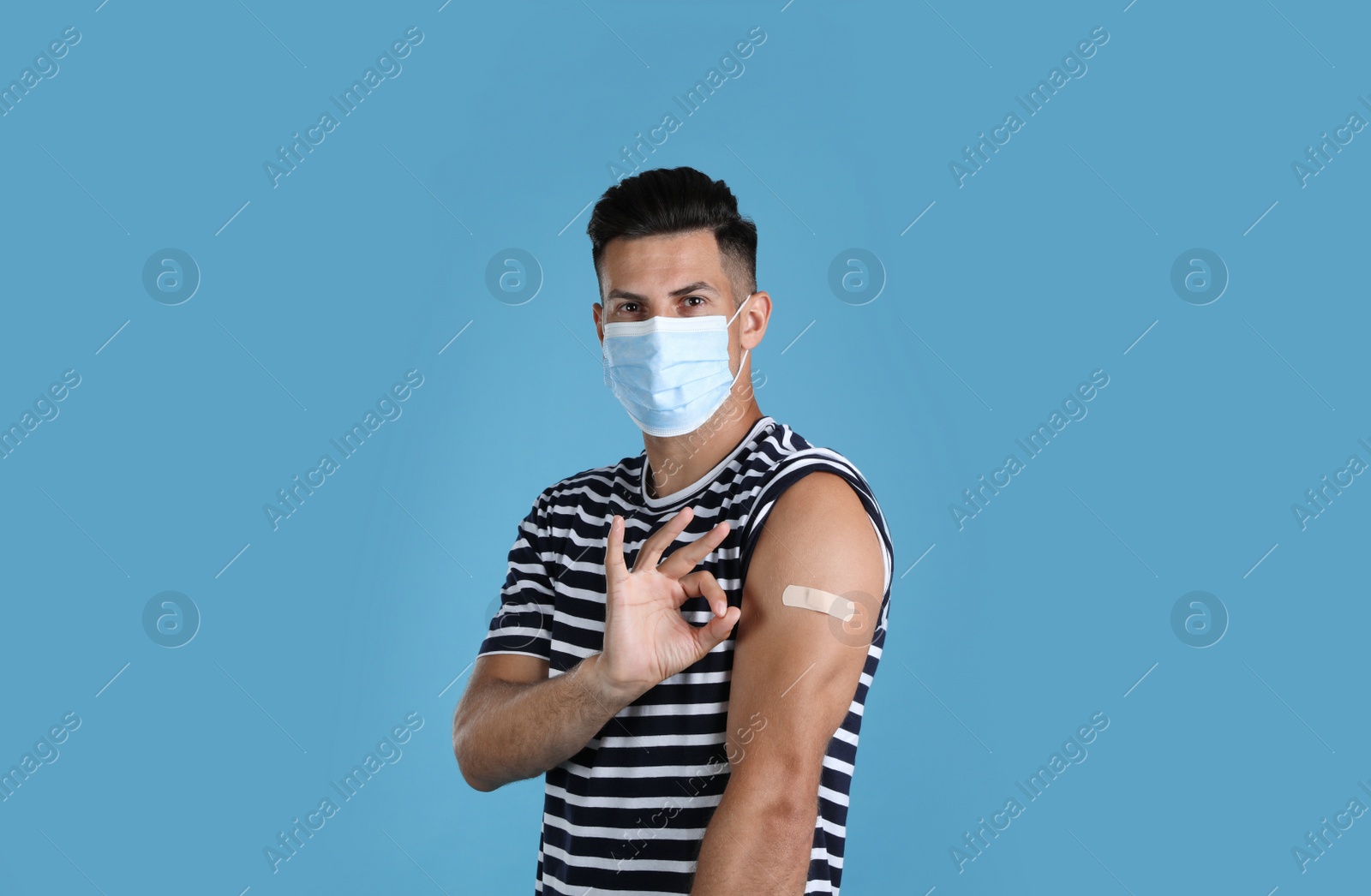 Photo of Vaccinated man with protective mask and medical plaster on his arm showing okay gesture against light blue background