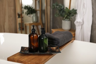 Wooden bath tray with bottles of shower gels and towel on tub indoors, space for text