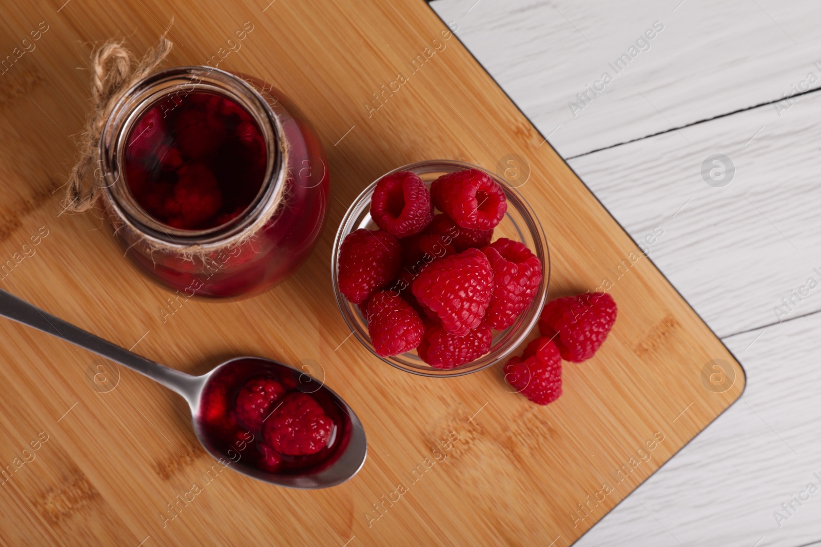 Photo of Jar of tasty canned raspberry jam and fresh berries in glass bowl on white wooden table, flat lay