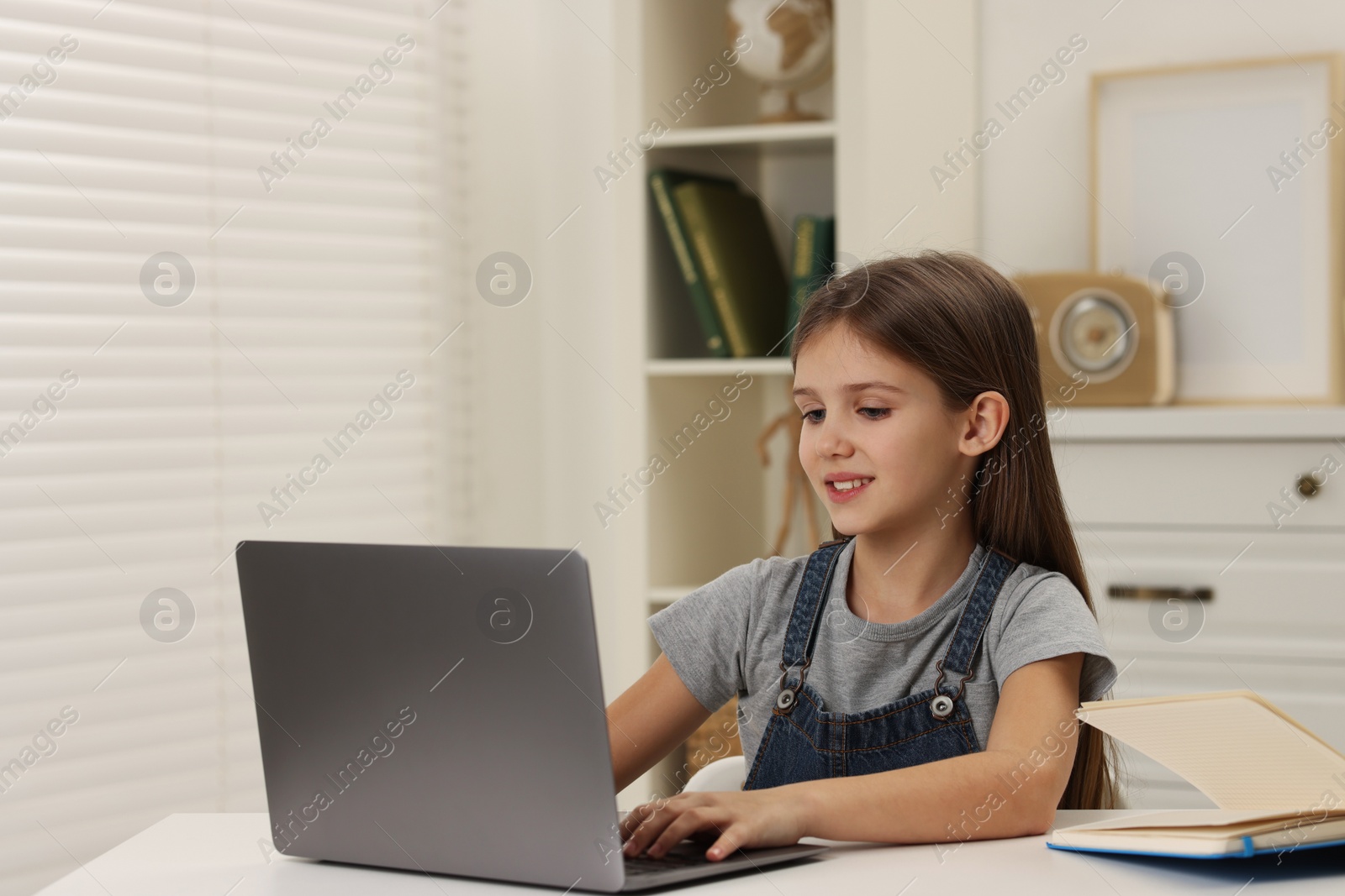 Photo of Cute girl using laptop at white table indoors