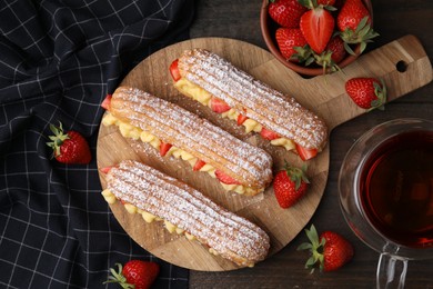 Photo of Delicious eclairs filled with cream, strawberries and tea on wooden table, top view