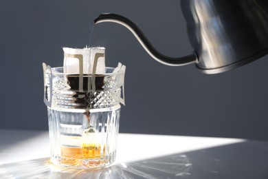 Photo of Pouring hot water into glass with drip coffee bag from kettle on light grey table, closeup