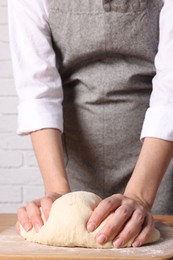 Photo of Woman kneading dough at wooden table near white brick wall, closeup