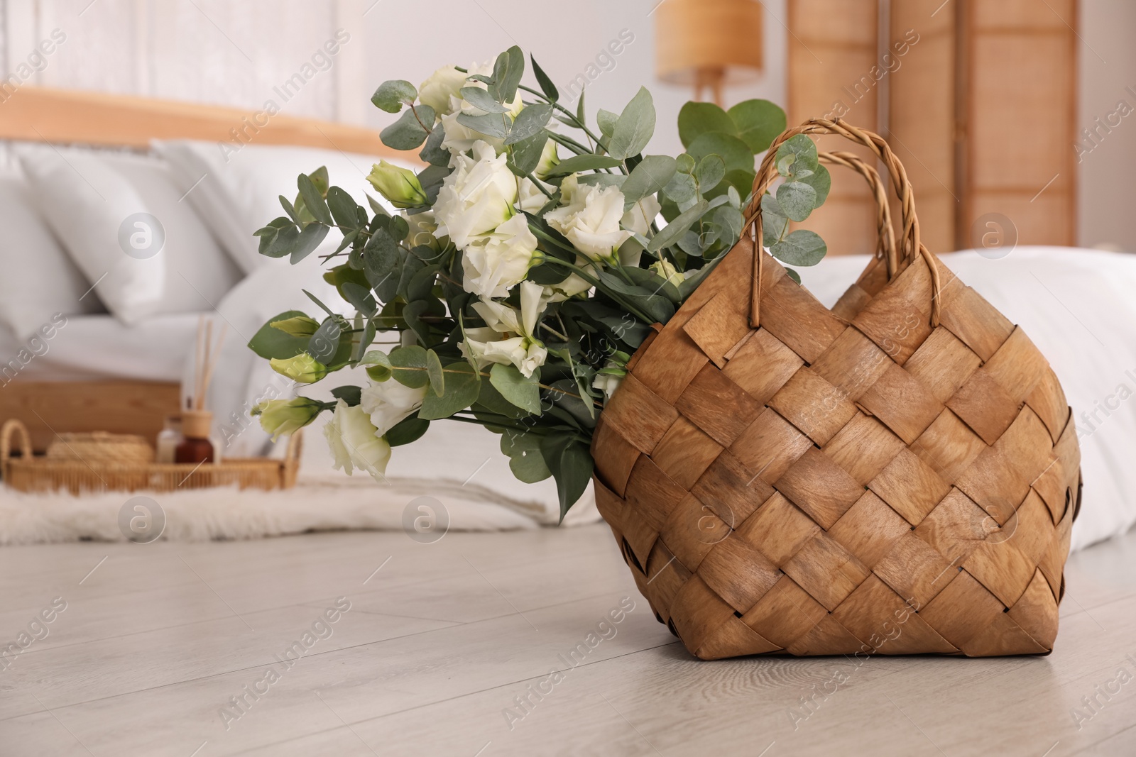 Photo of Stylish wicker basket with fresh eucalyptus branches and flowers on floor in bedroom