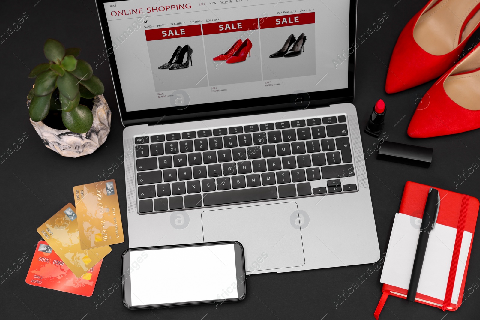 Photo of Online store website on laptop screen. Computer, smartphone, credit cards, women's shoes, stationery and lipstick on black background, above view