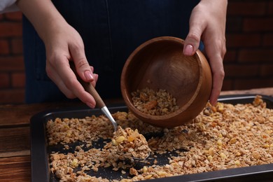 Woman putting granola from baking tray into bowl at table, closeup