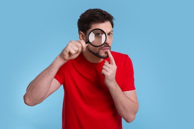 Confused man looking through magnifier glass on light blue background