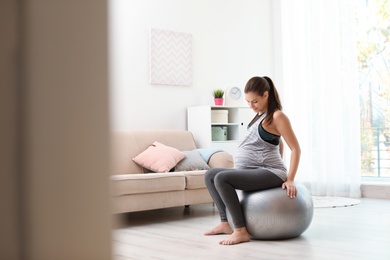 Photo of Young pregnant woman in fitness clothes sitting on exercise ball at home. Space for text
