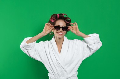 Beautiful young woman in bathrobe with hair curlers and sunglasses on green background