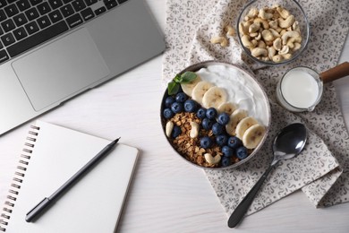 Delicious granola in bowl, stationery and laptop on white wooden table, flat lay