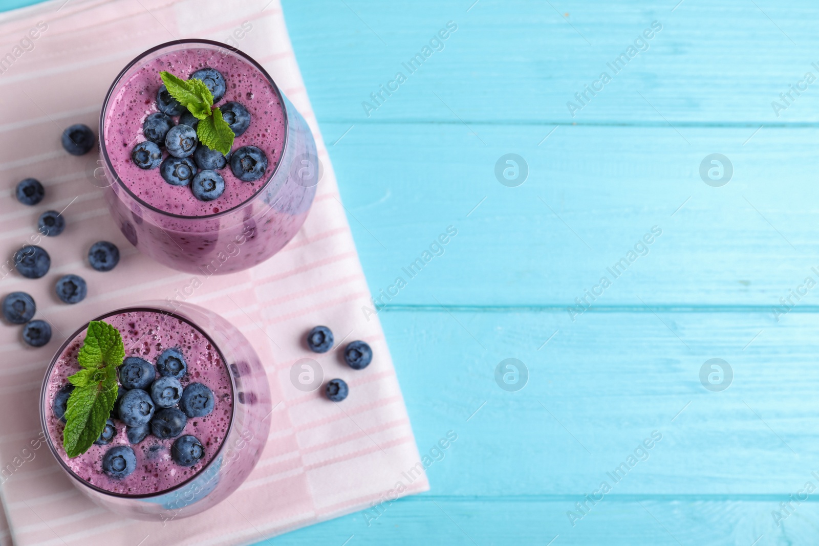 Photo of Glasses of tasty blueberry smoothie and fabric on light blue wooden table, flat lay. Space for text