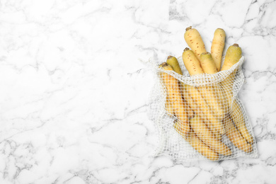 Many raw white carrots in mesh bag on marble table, top view. Space for text