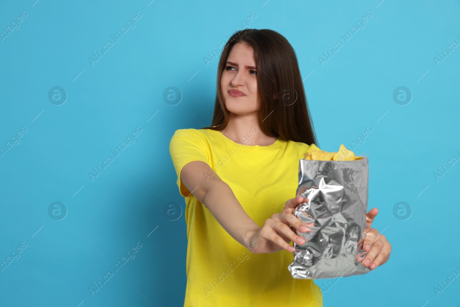 Photo of Unhappy young woman with bag of potato chips on light blue background