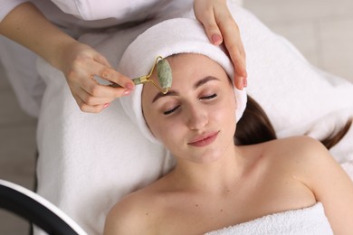 Photo of Cosmetologist making face massage with roller to client in clinic, top view