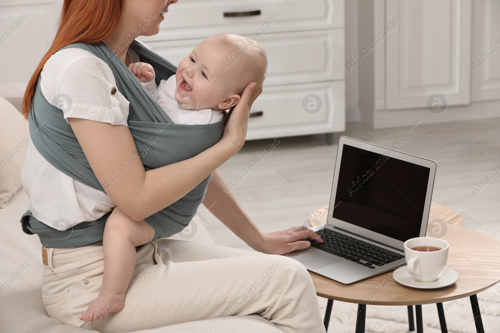 Photo of Mother using laptop while holding her child in sling (baby carrier) at home