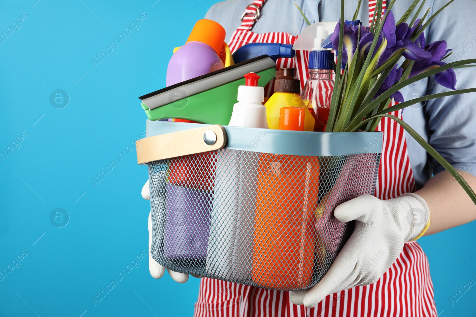 Photo of Woman holding basket with spring flowers and cleaning supplies on light blue background, closeup