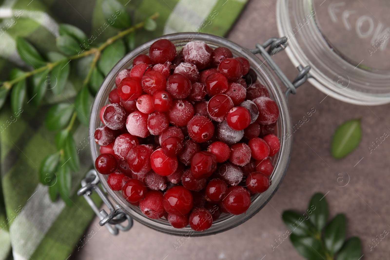 Photo of Frozen red cranberries in glass jar and green leaves on brown textured table, top view