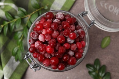 Frozen red cranberries in glass jar and green leaves on brown textured table, top view