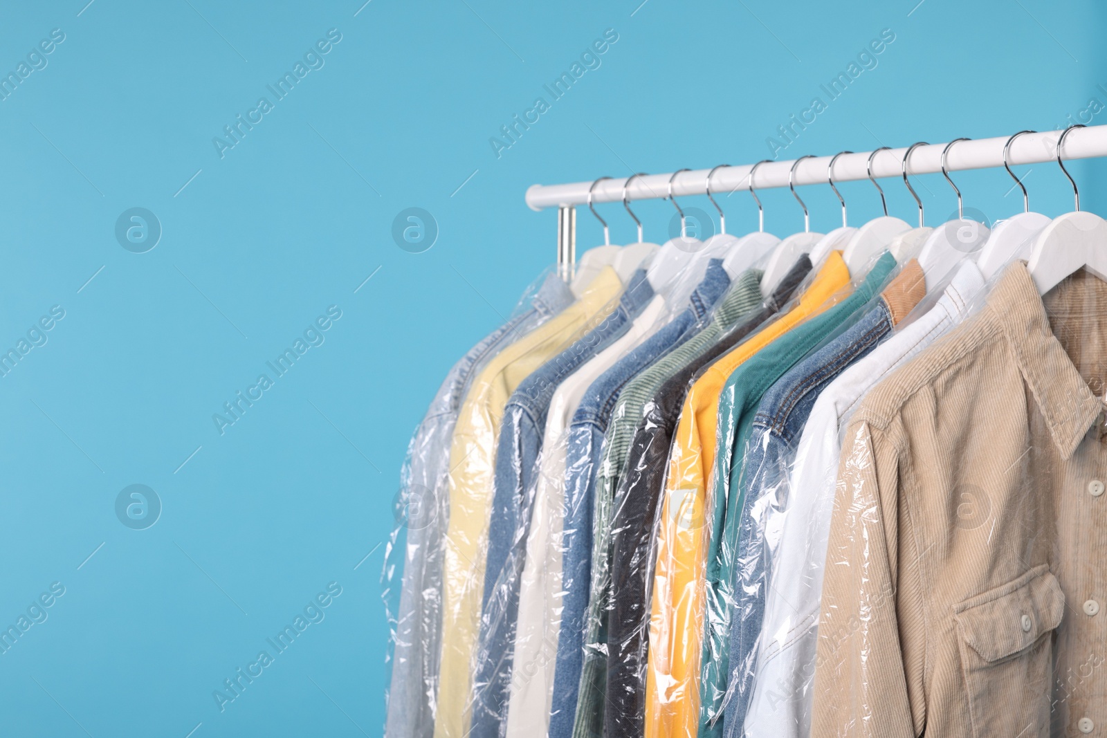 Photo of Dry-cleaning service. Many different clothes in plastic bags hanging on rack against light blue background, space for text