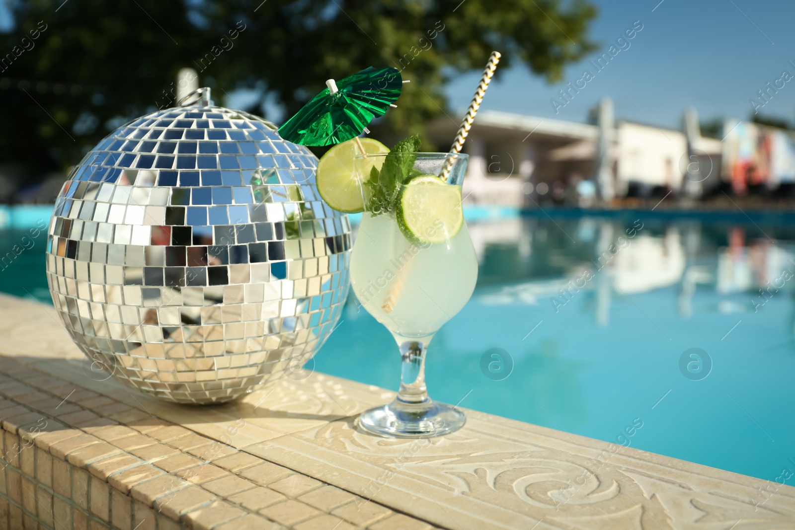 Photo of Tasty refreshing cocktail and shiny disco ball on edge of swimming pool. Party items